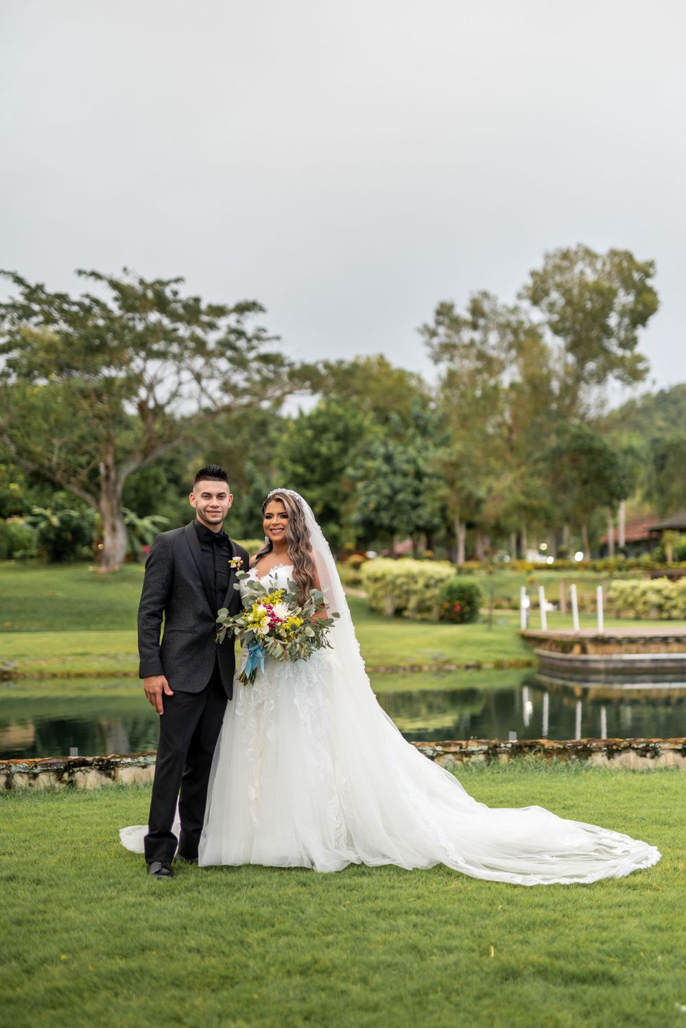 Newlyweds stand together on manicured lawn with a scenic pond backdrop.