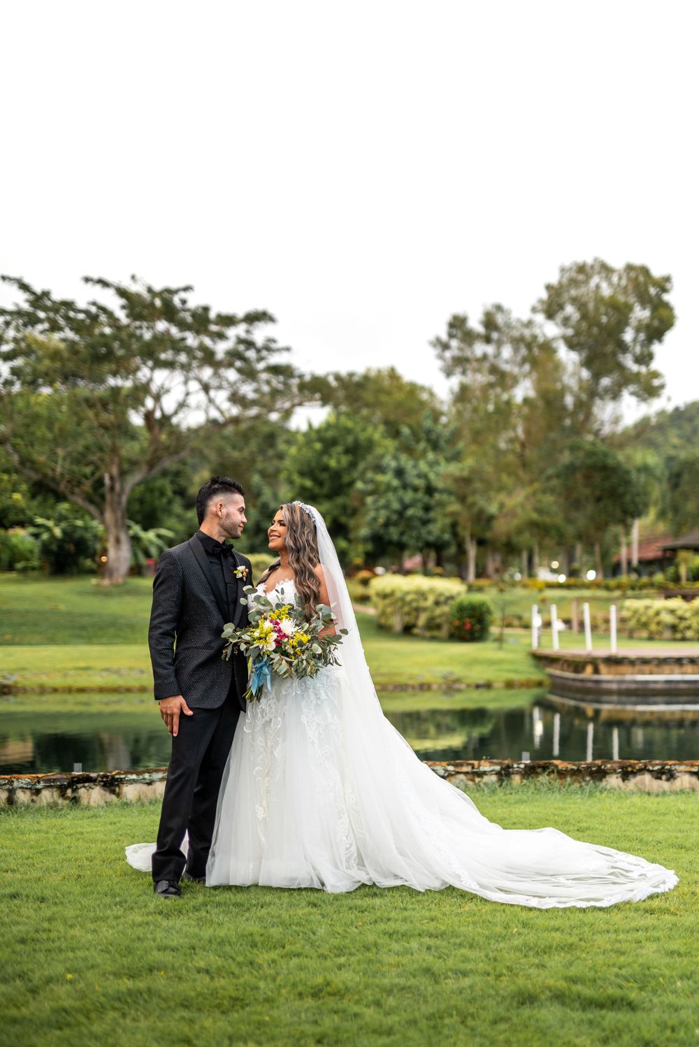 Wedding couple shares a lighthearted moment on lush grounds with pond view.