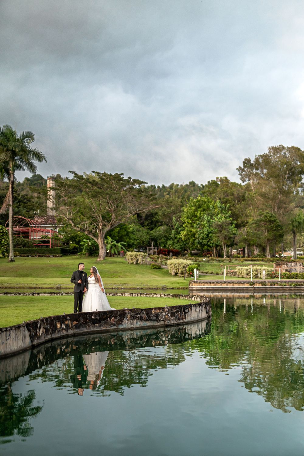 Wedding couple stands on bridge over reflective pond surrounded by tropical landscaping.