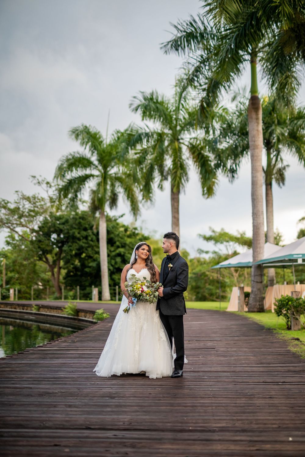 Wedding couple poses on wooden boardwalk with towering palm trees overhead.