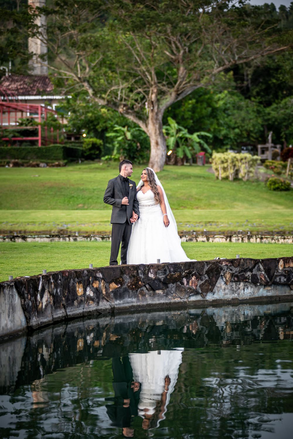 A couple stands on a stone wall reflecting in a tranquil pond surrounded by lush gardens and tropical greenery.