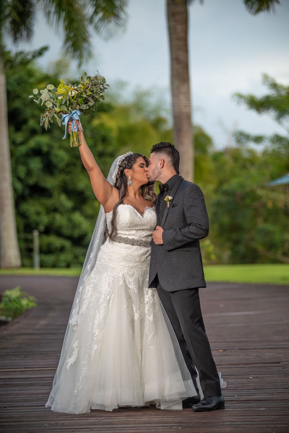 A couple celebrates with joy on a wooden deck with tropical foliage in the background.