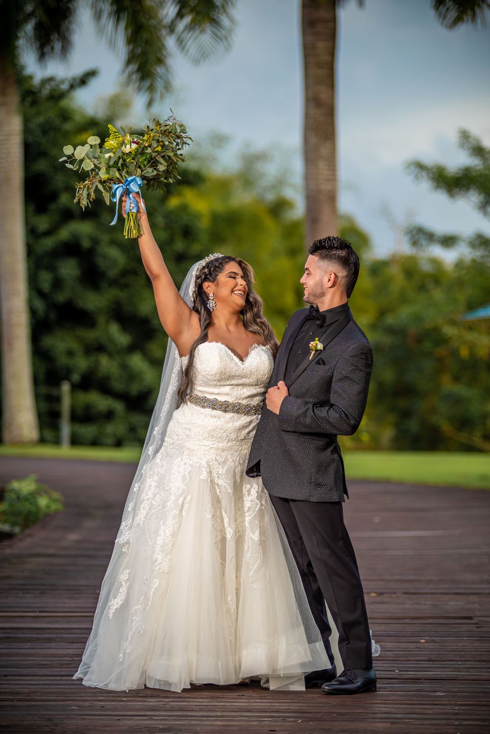 A couple shares an exuberant moment on a wooden boardwalk surrounded by palm trees.