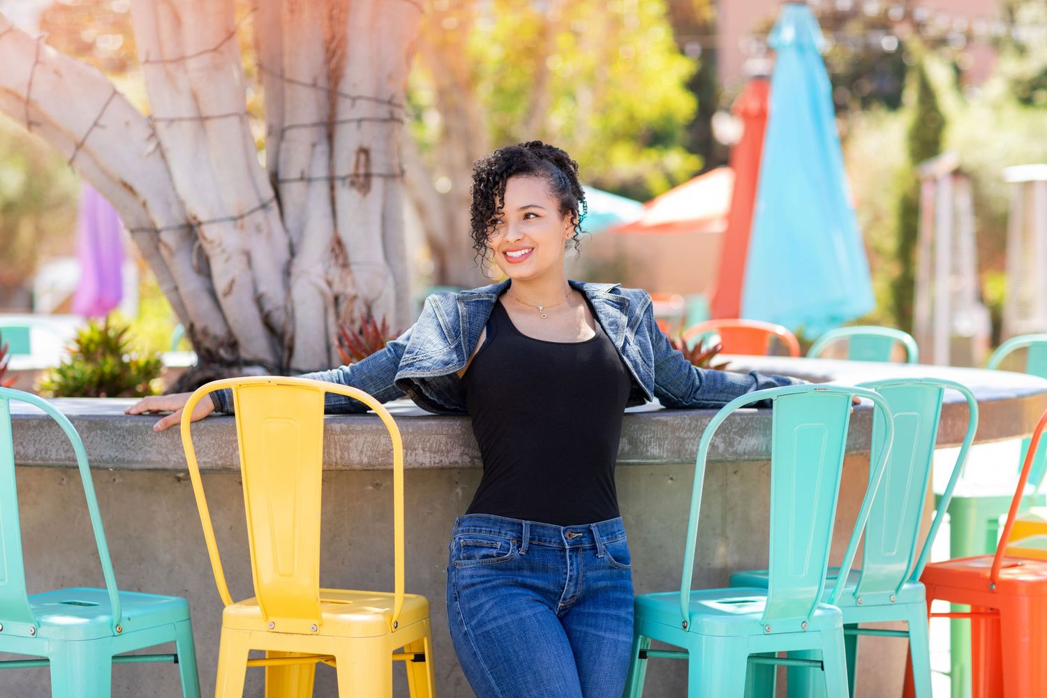 A high school senior poses outdoors on a patio with colorful metal chairs in turquoise and yellow on a sunny day.