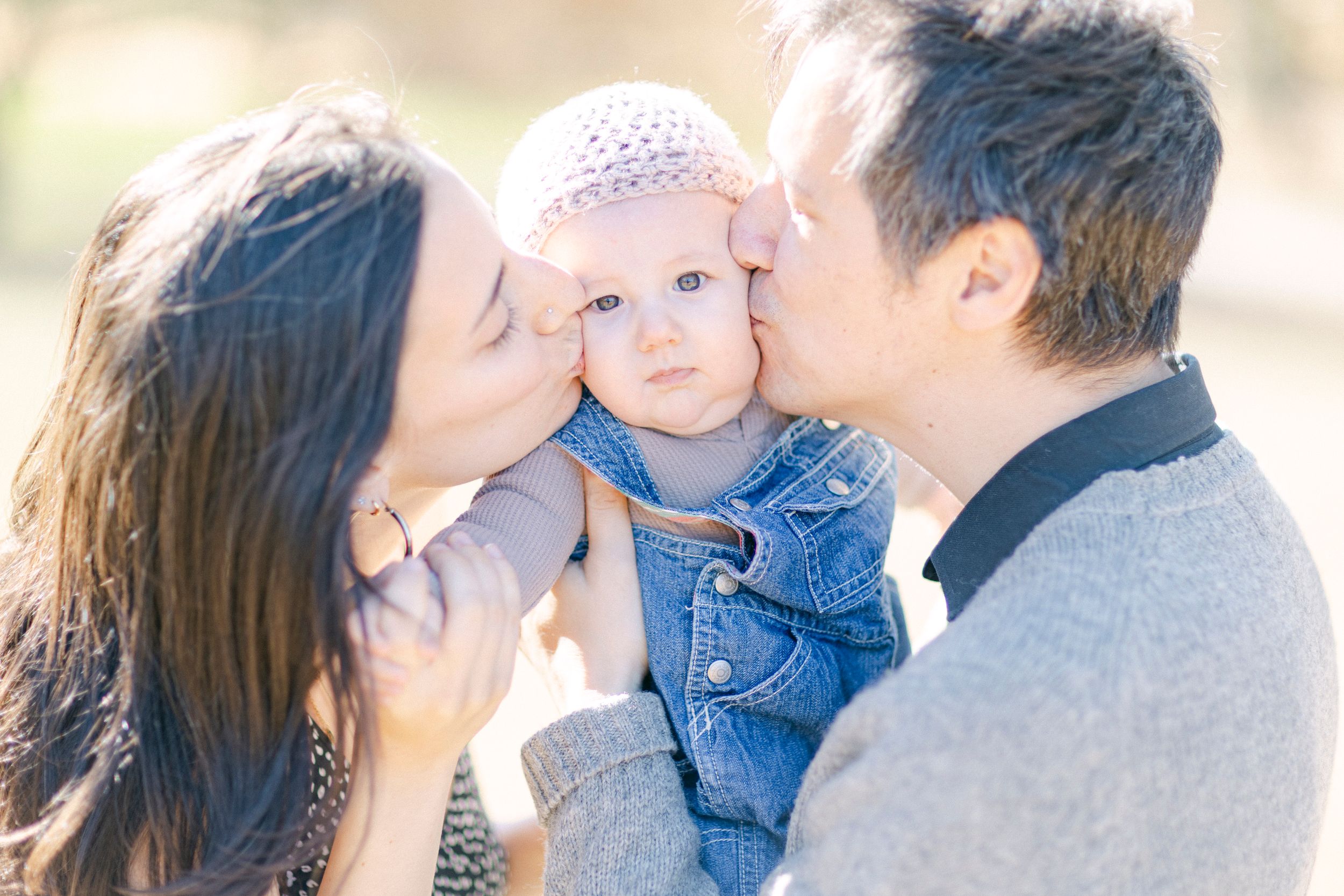 Parents giving their baby sweet kisses in a sun-drenched outdoor photo.