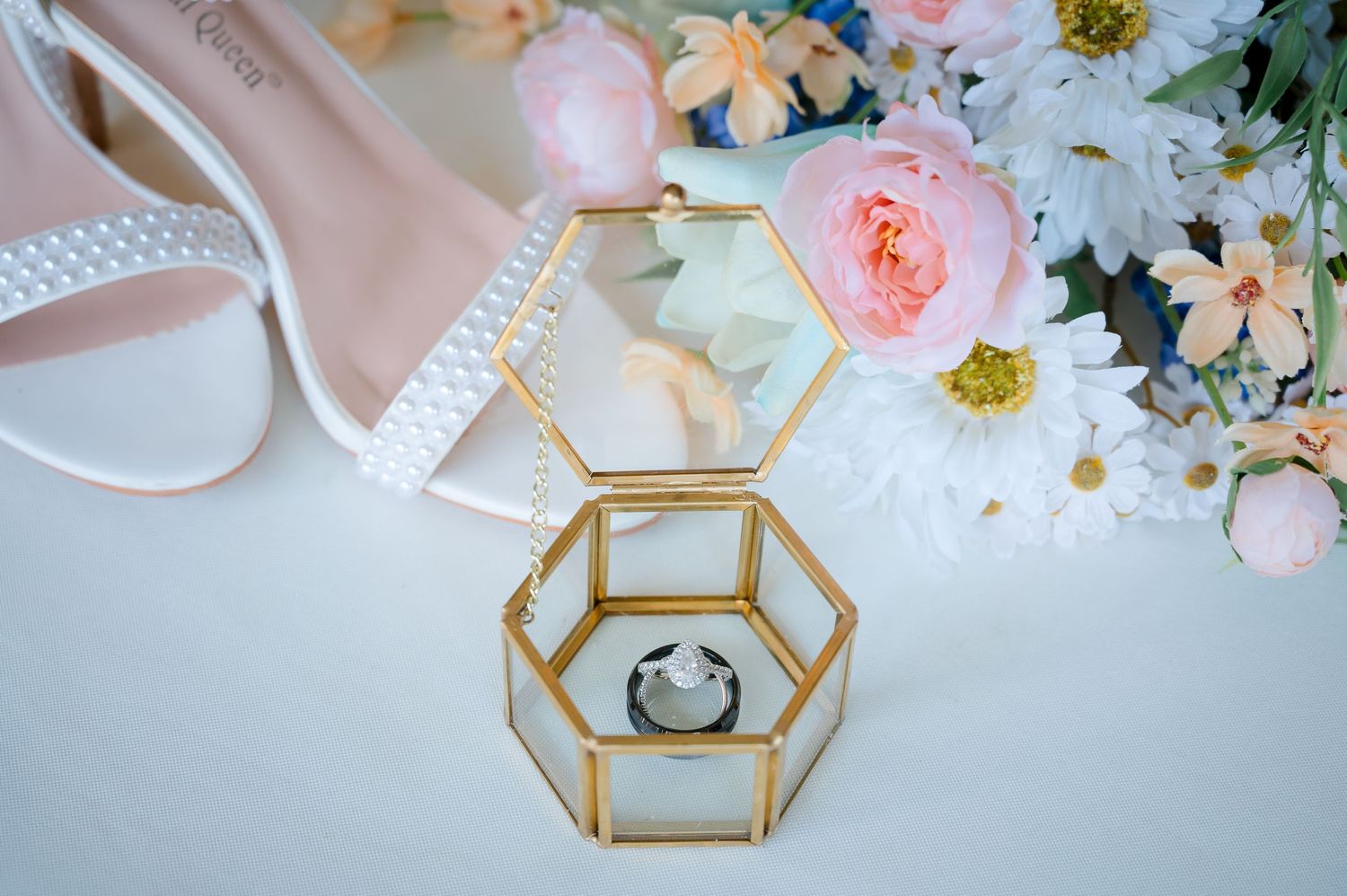 Wedding details featuring white shoes, geometric ring box, and delicate pink floral arrangements.