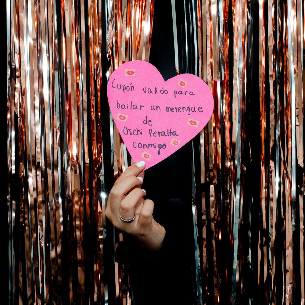 A hand holding a pink heart-shaped note against a sparkly curtain backdrop.