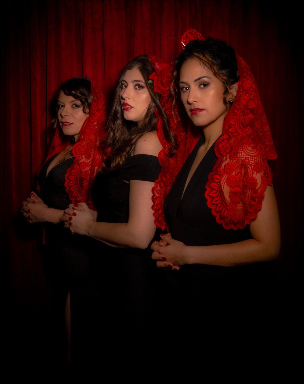 Three performers in black dresses pose dramatically against a deep red backdrop in a theatrical sequence.