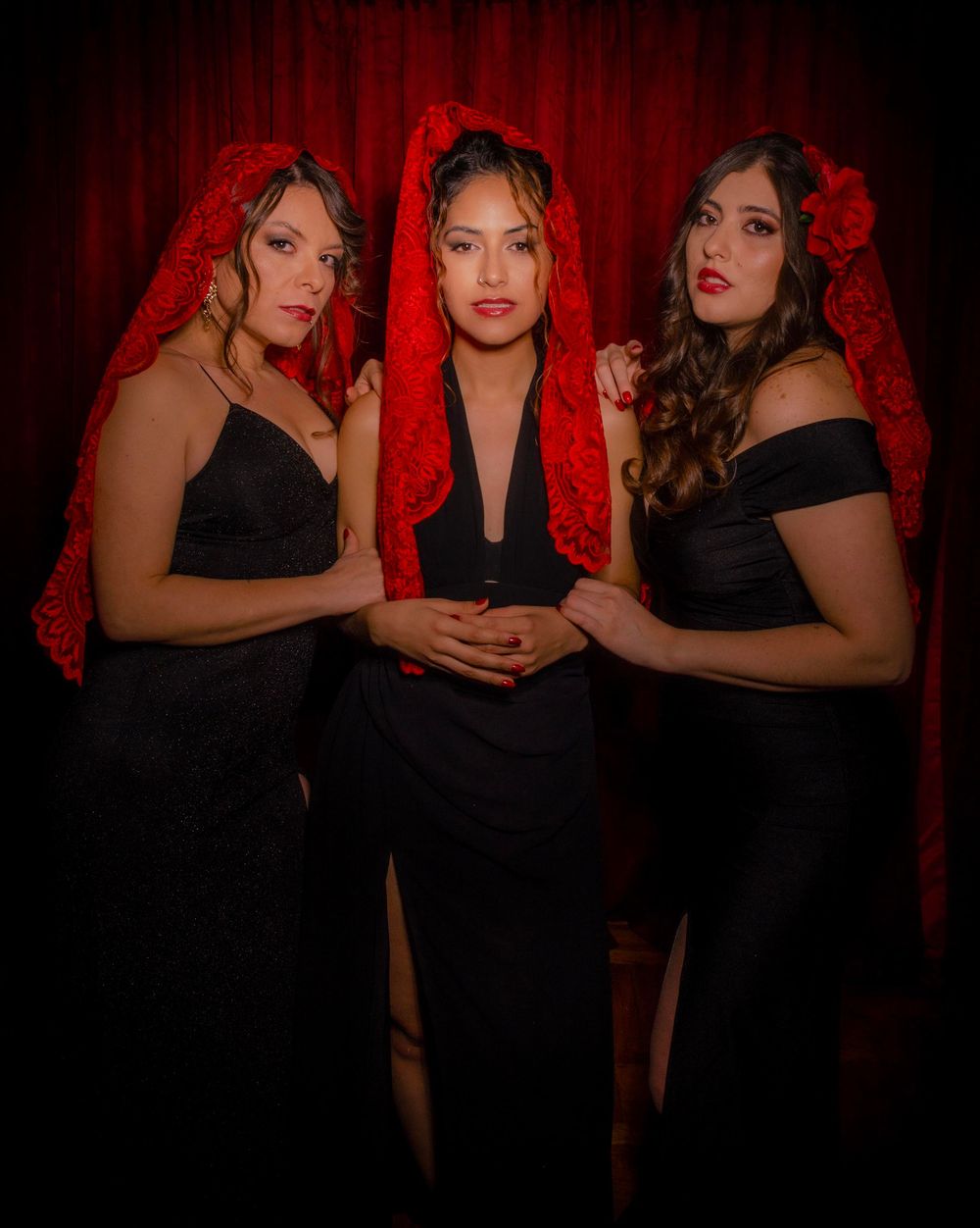 Three people in black dresses pose together against a dramatic red backdrop in a glamorous photo session.