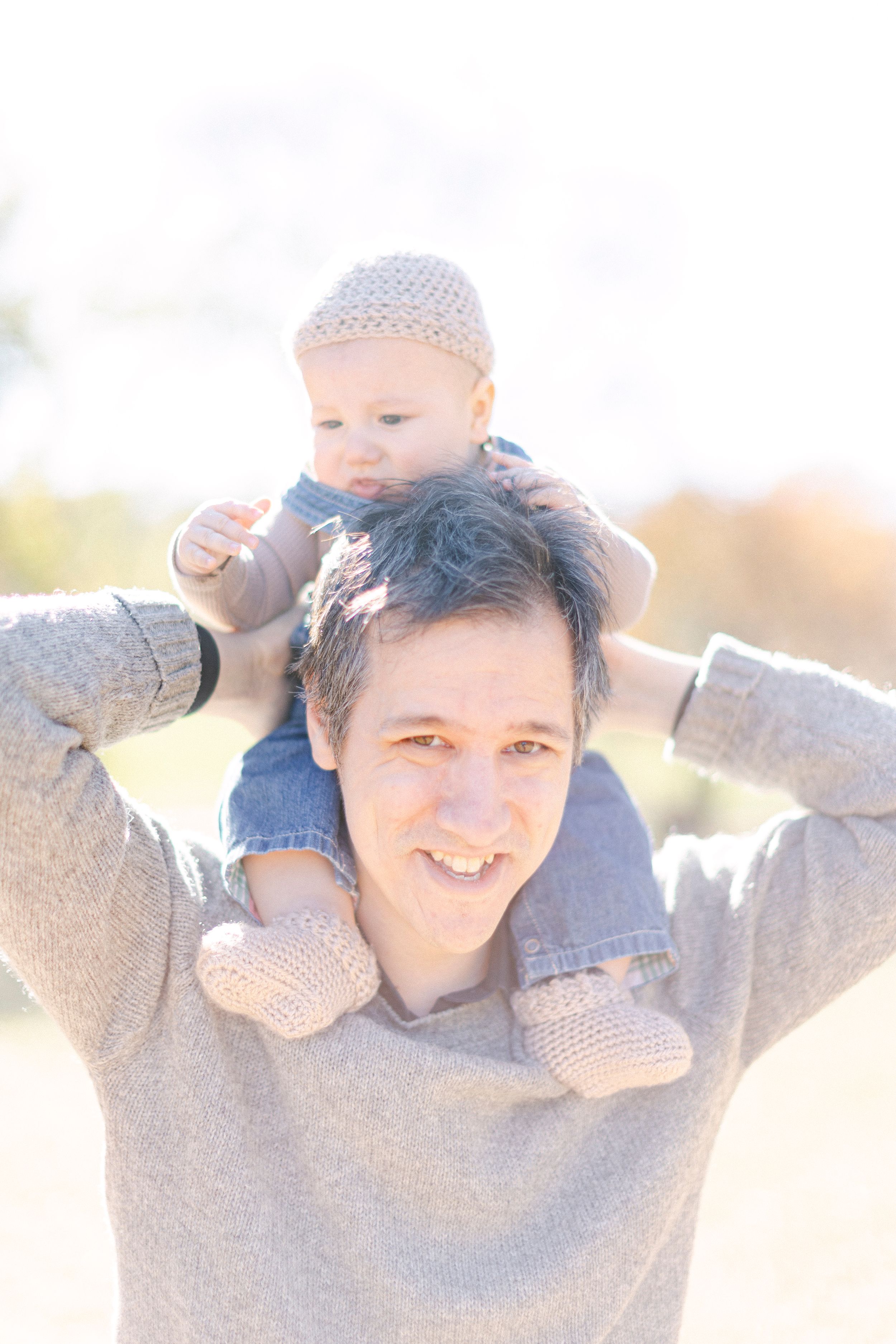 A baby wearing a knit hat sits on shoulders during a bright outdoor photo session.