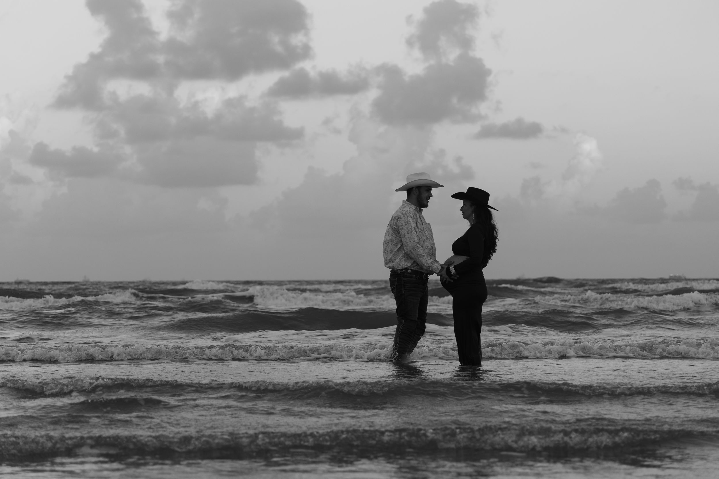 A couple stands silhouetted against dramatic ocean waves and cloudy skies in a black and white photograph.