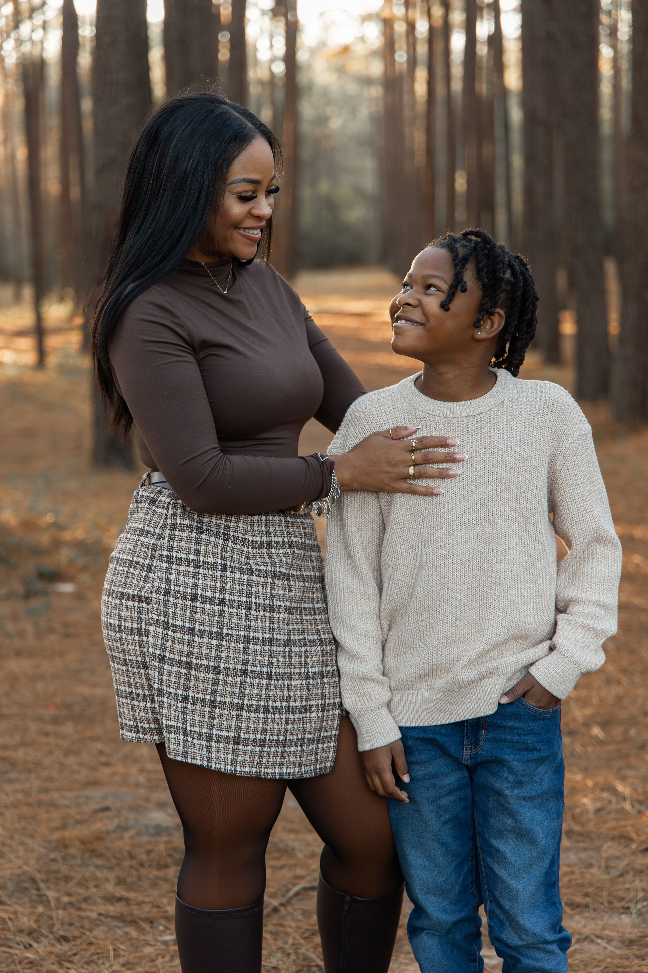 A heartwarming outdoor family photoshoot in a sunlit pine forest with autumn leaves on the ground.