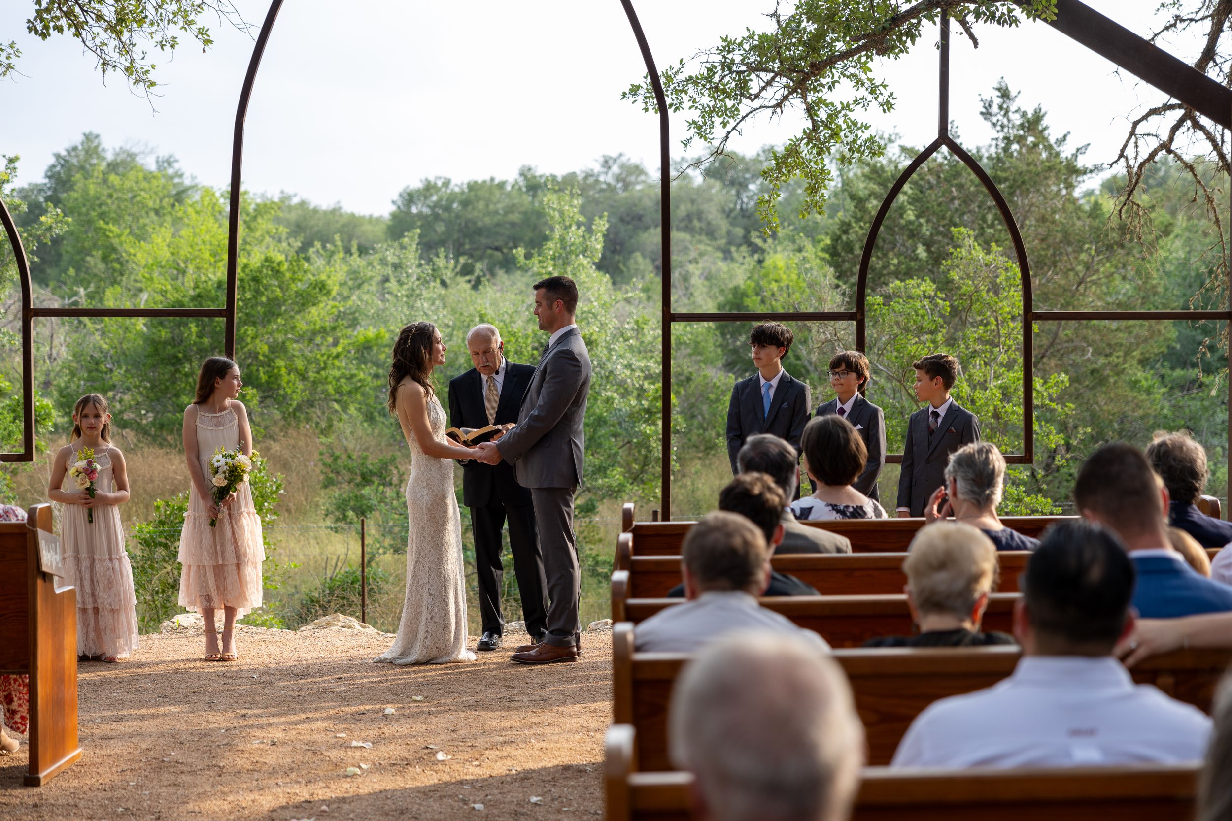 An outdoor wedding ceremony setup with wooden benches and natural tree canopy.