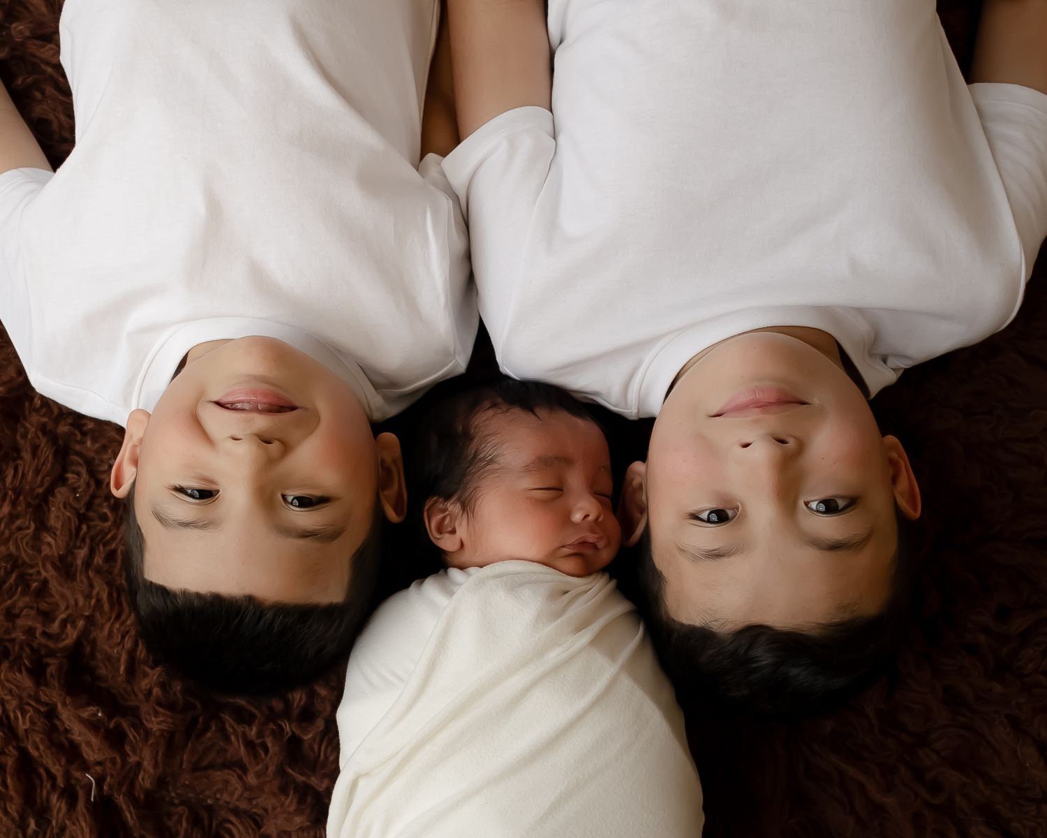 Overhead view of three family members lying on brown fabric with center infant wrapped in white.