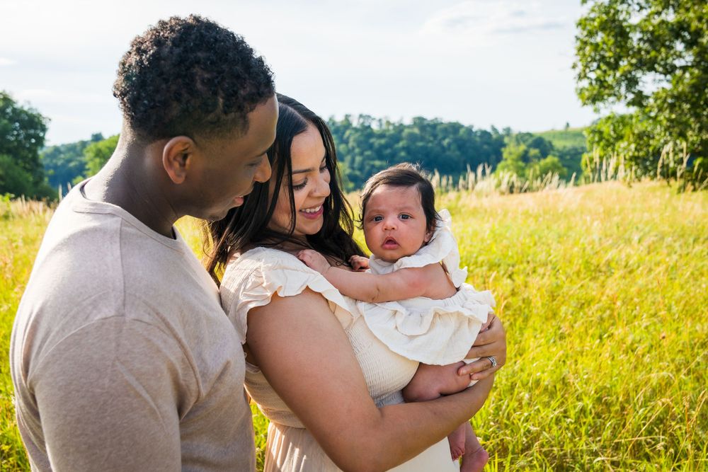 Parents lovingly gaze at their baby while standing in tall grass field during sunset.