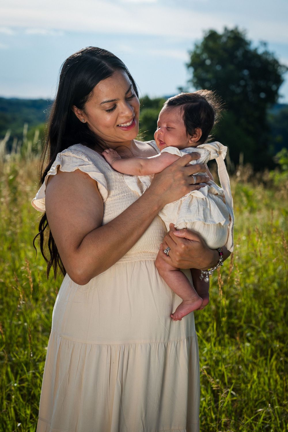 Mother in cream dress holds and smiles at baby during outdoor photoshoot.