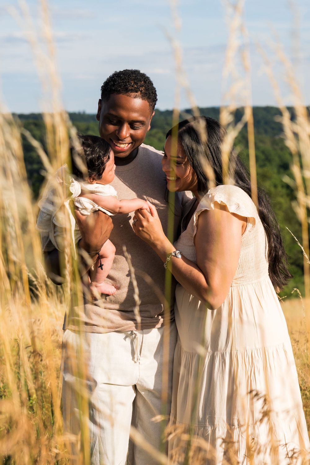 Parents share intimate moment with newborn in tall grass field at sunset.