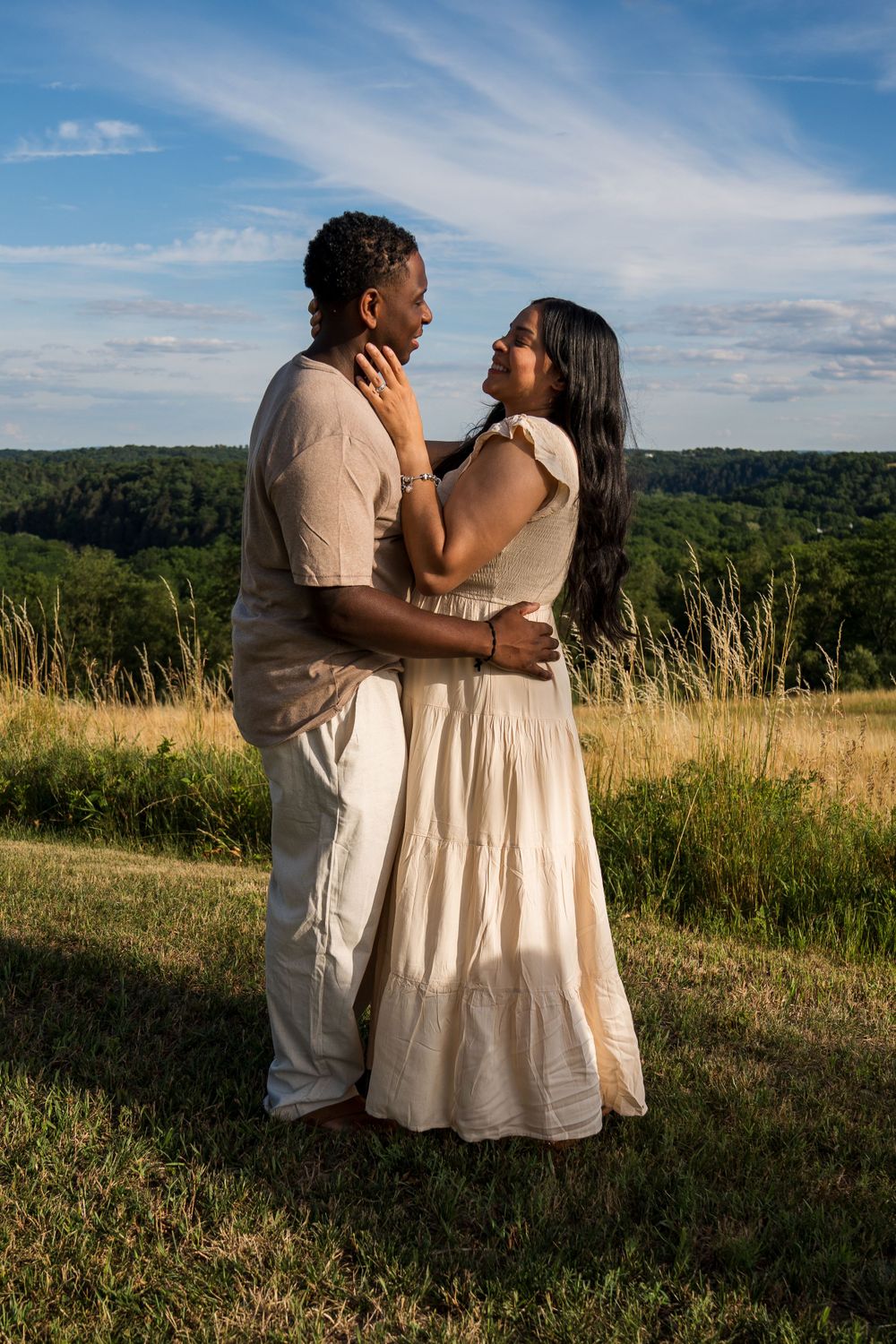 A couple embraces in a romantic moment while standing in a hilltop meadow during sunset.