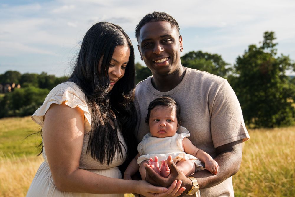A couple holds their newborn baby while standing in a sunny field with trees in the background.
