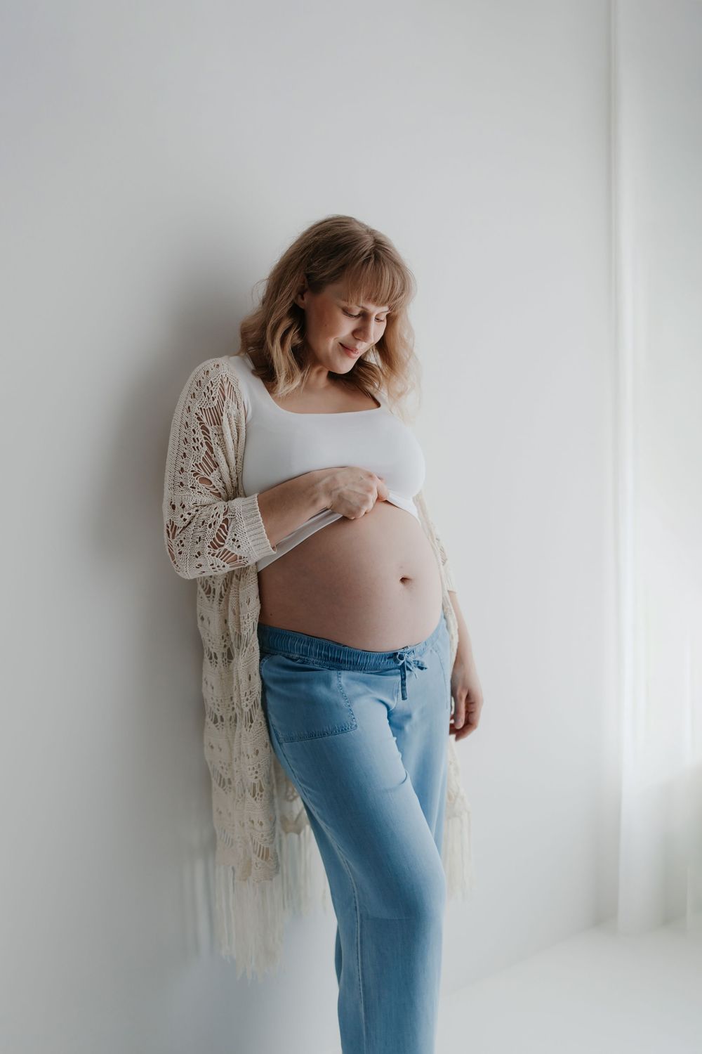 Pregnant person in blue pants and white top poses against white wall in maternity photo series.