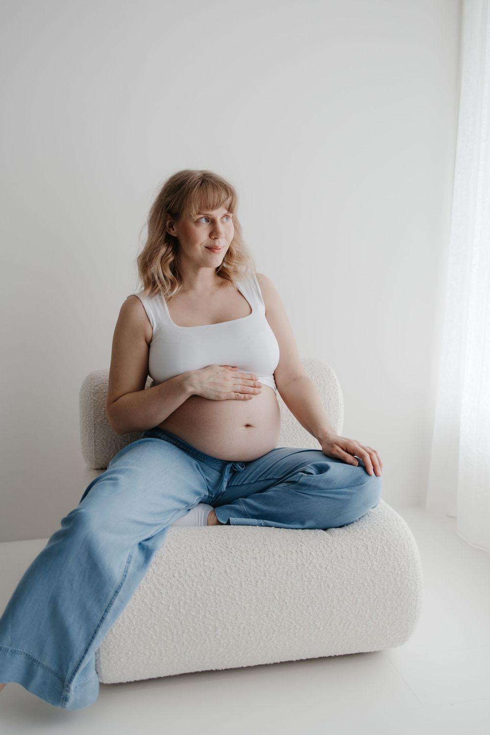 Minimalist maternity photo series showing pregnant woman in white tank top and jeans reclining on light couch.