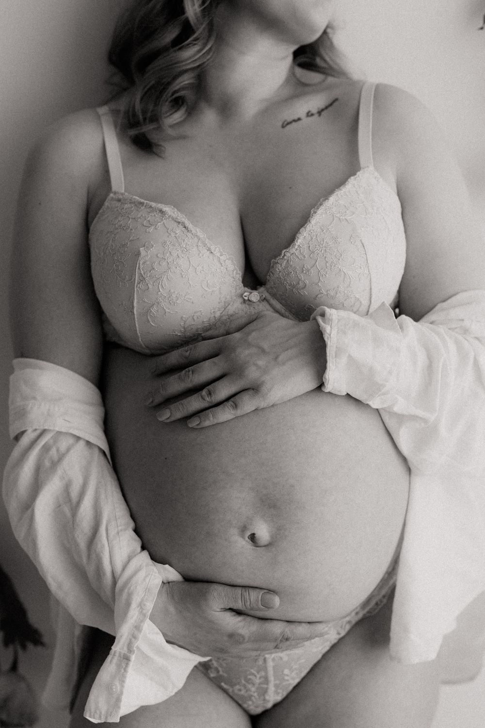 Black and white maternity photo of pregnant woman in lace lingerie cradling baby bump with open white shirt.