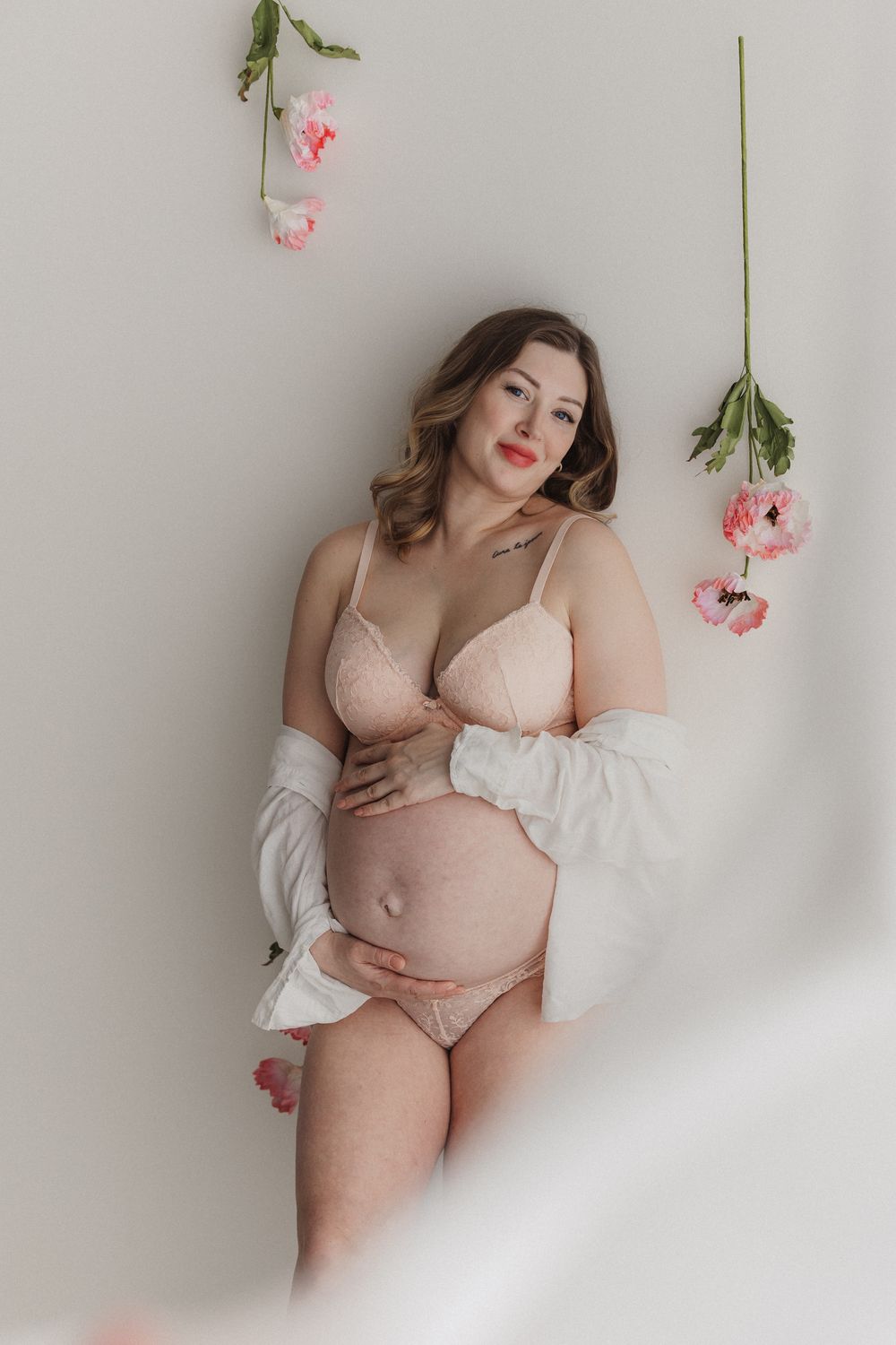 Pregnant woman in blush lingerie and white robe cradling baby bump, surrounded by hanging pink flowers.