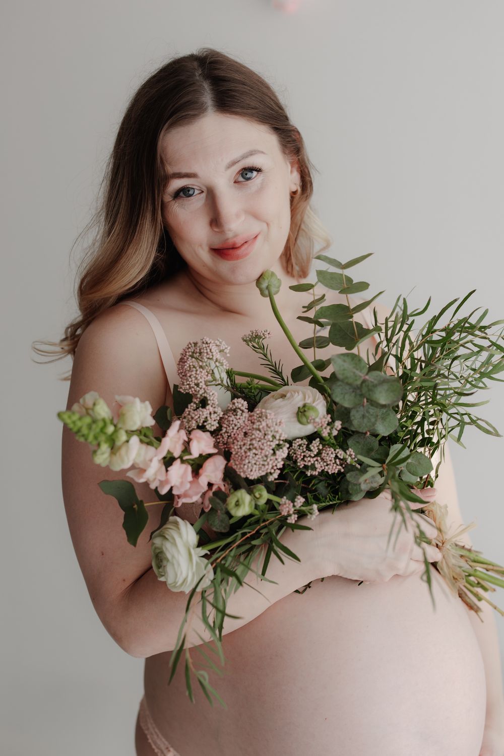 Pregnant woman in pink dress holding a bouquet of pink and white flowers, smiling softly at the camera.