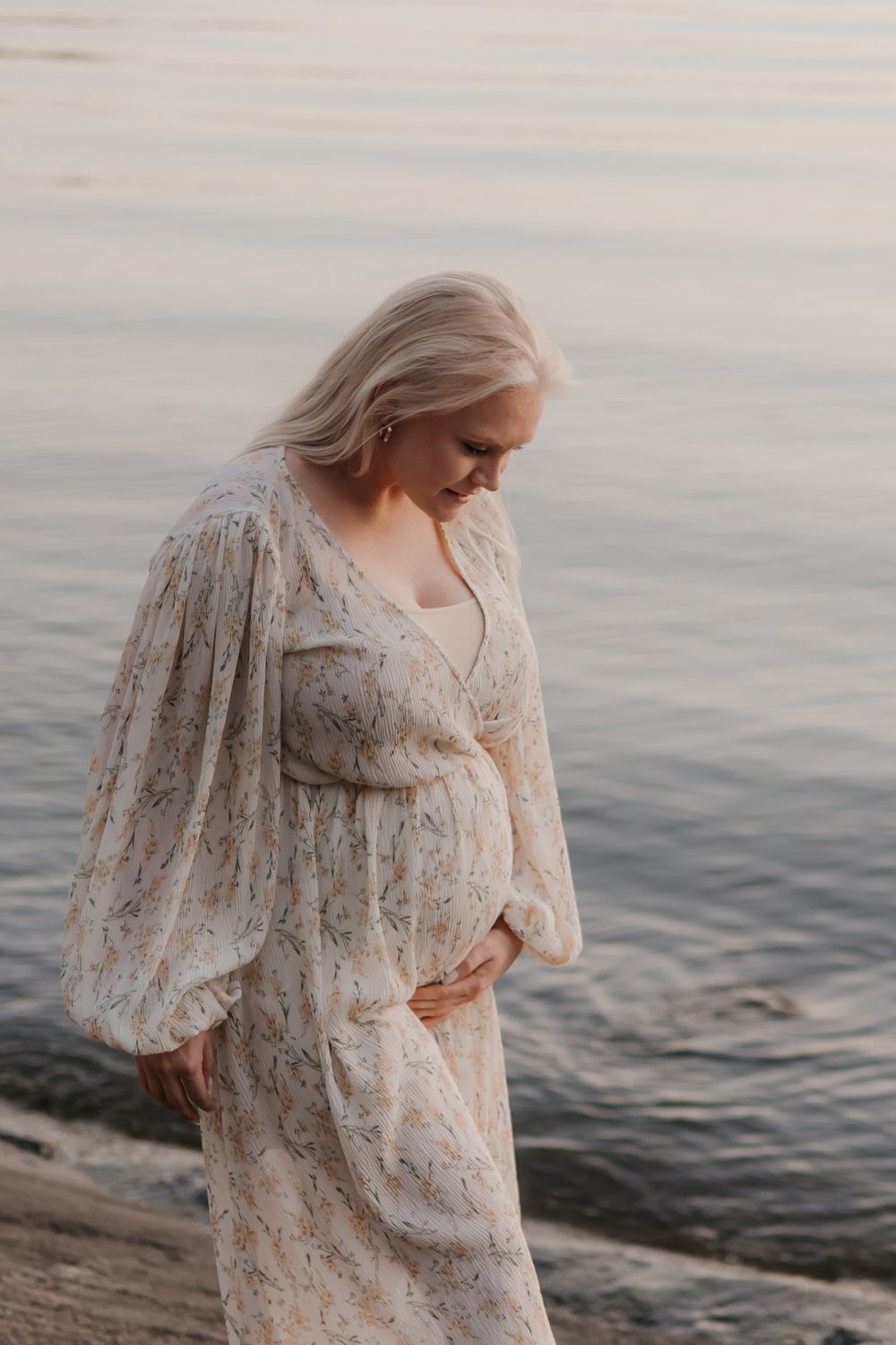 Person in vintage-style floral dress poses by calm water at dusk.