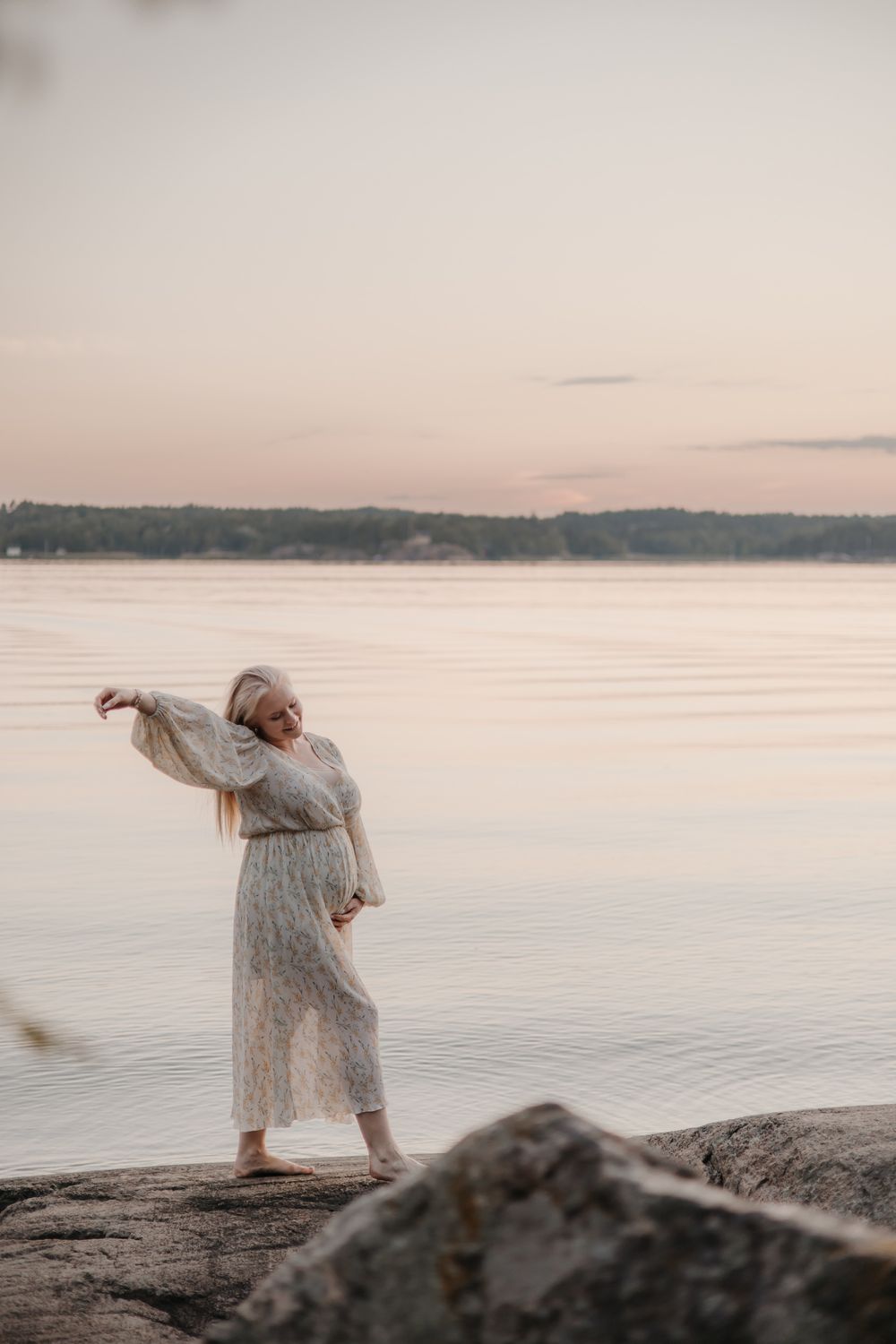 Person in flowing dress poses dramatically on rocky shoreline at sunset.