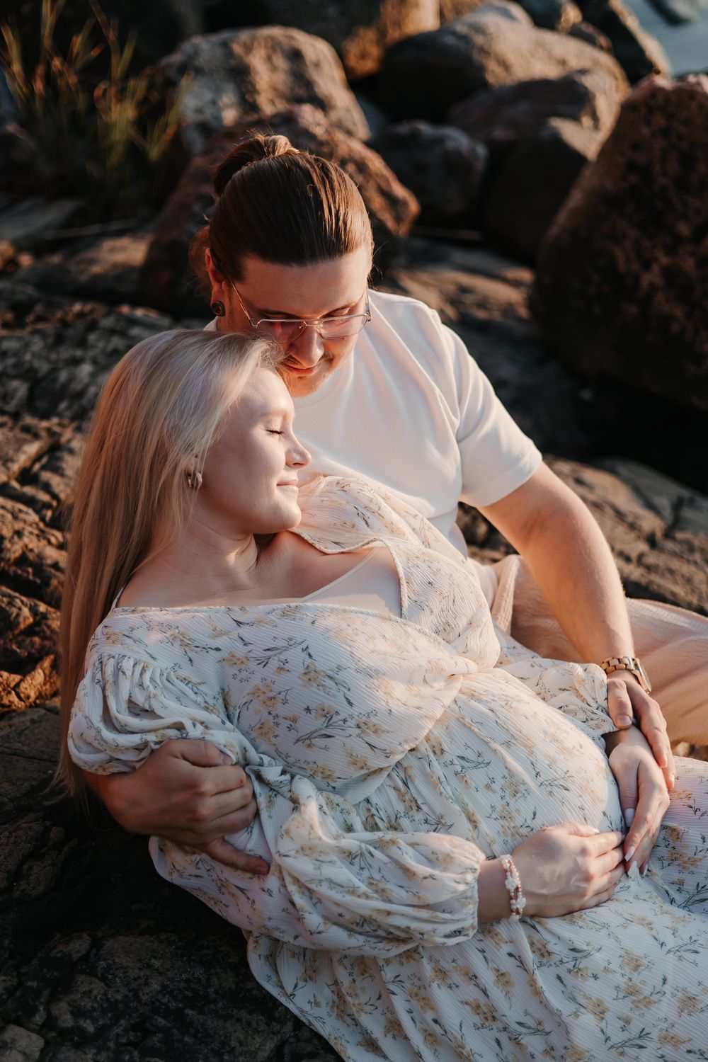 Couple in white clothing share intimate moment on rocky beach during golden hour.