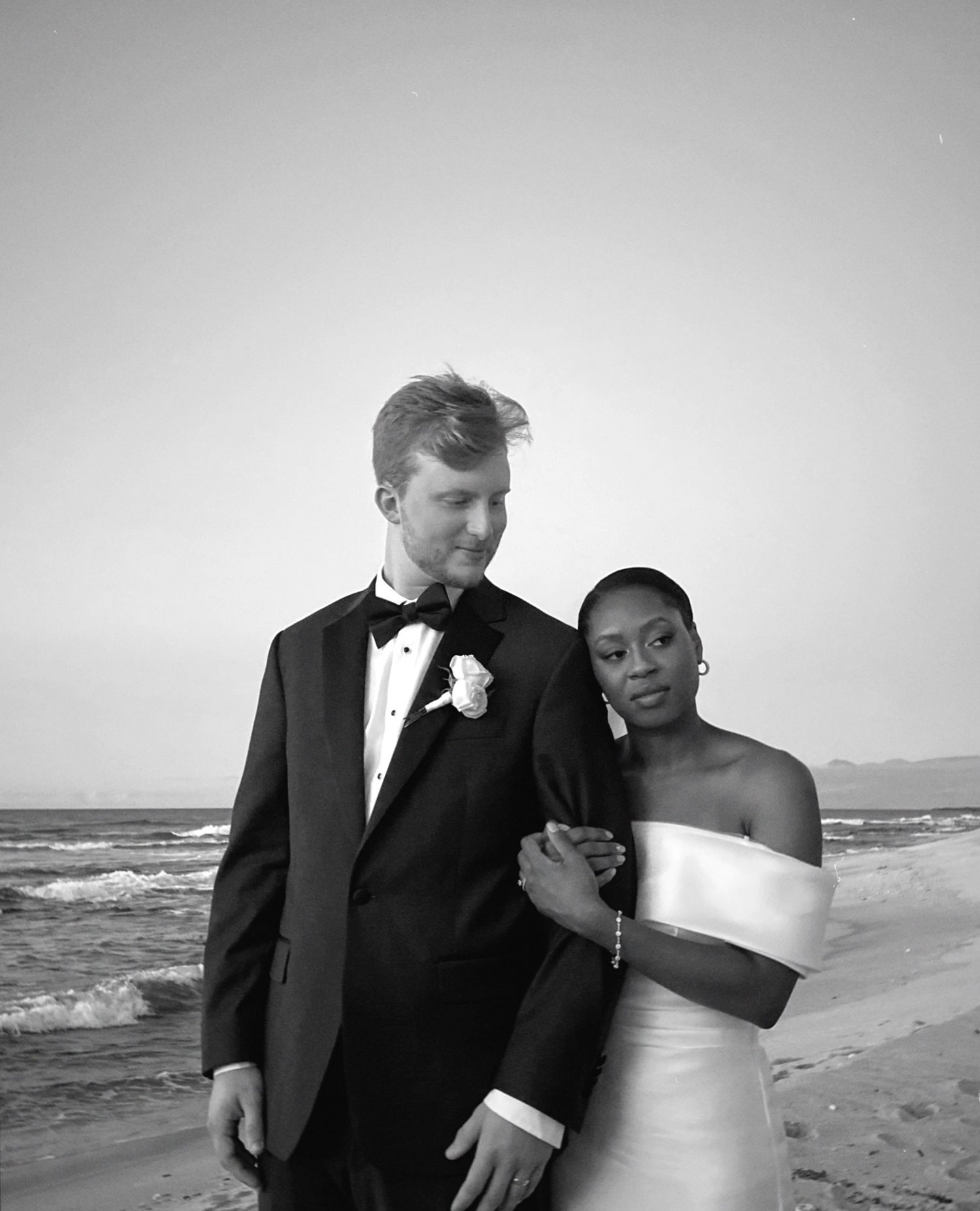 A couple in formal wedding attire poses together on a beach at sunset in black and white photography.