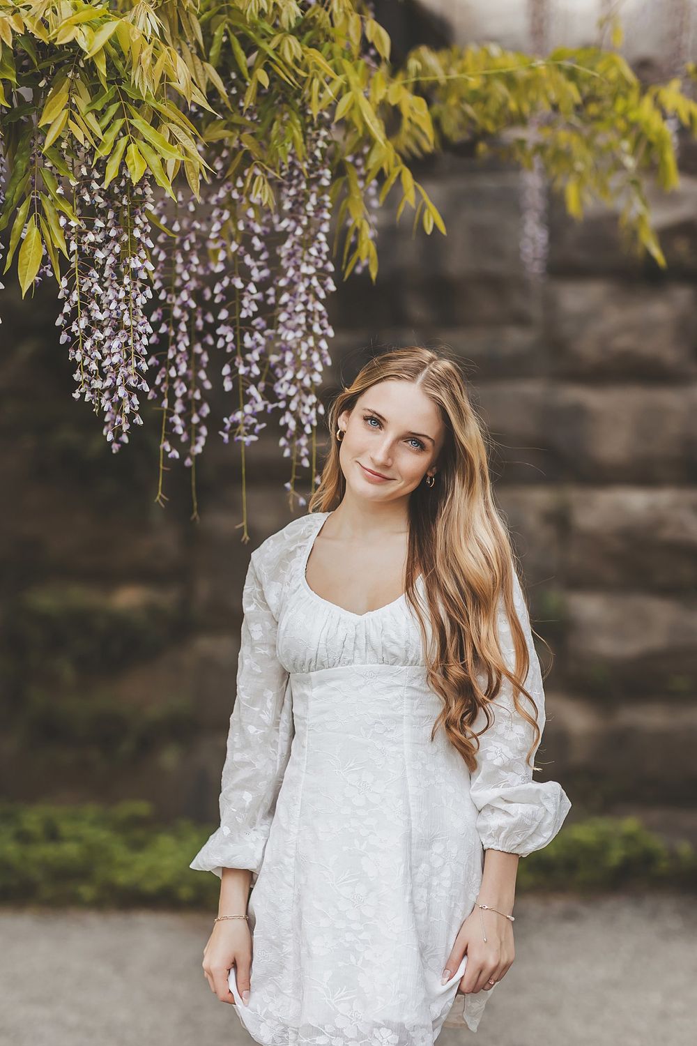 Spring portrait session under blooming wisteria with stone wall backdrop.