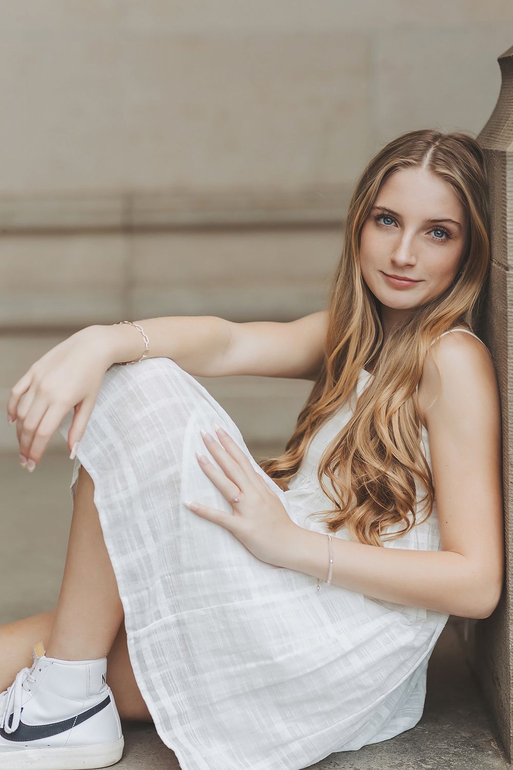 Portrait of a woman in white dress and sneakers sitting casually against a concrete wall.