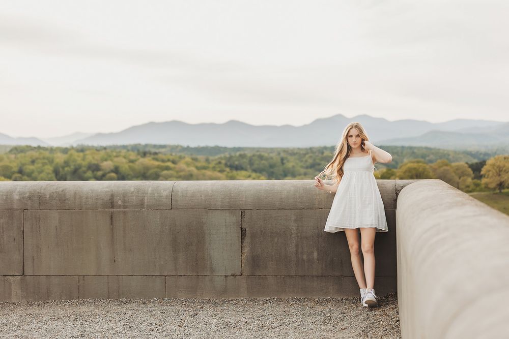Woman in white dress and sneakers poses along concrete wall with mountain vista.
