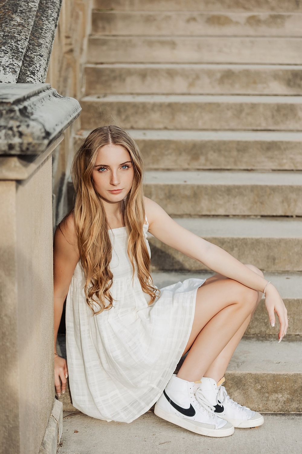 Woman in white dress and Nike sneakers poses elegantly on stone steps.