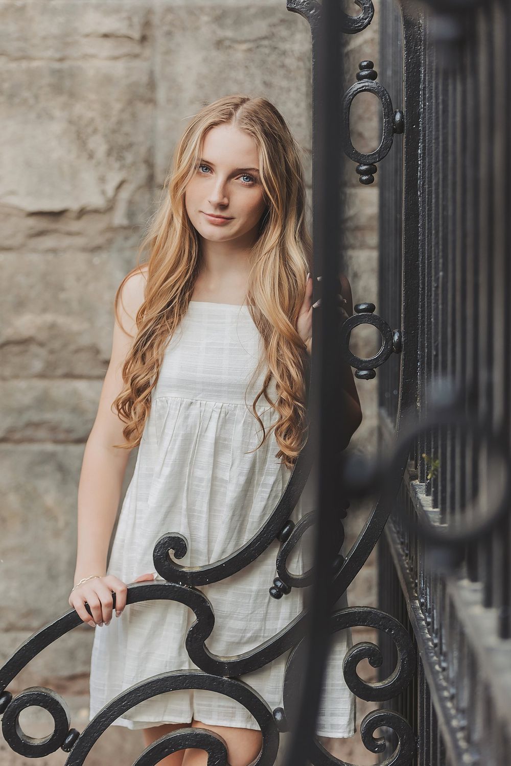 Long wavy hair and white dress complement the rustic stone wall and decorative iron gate.