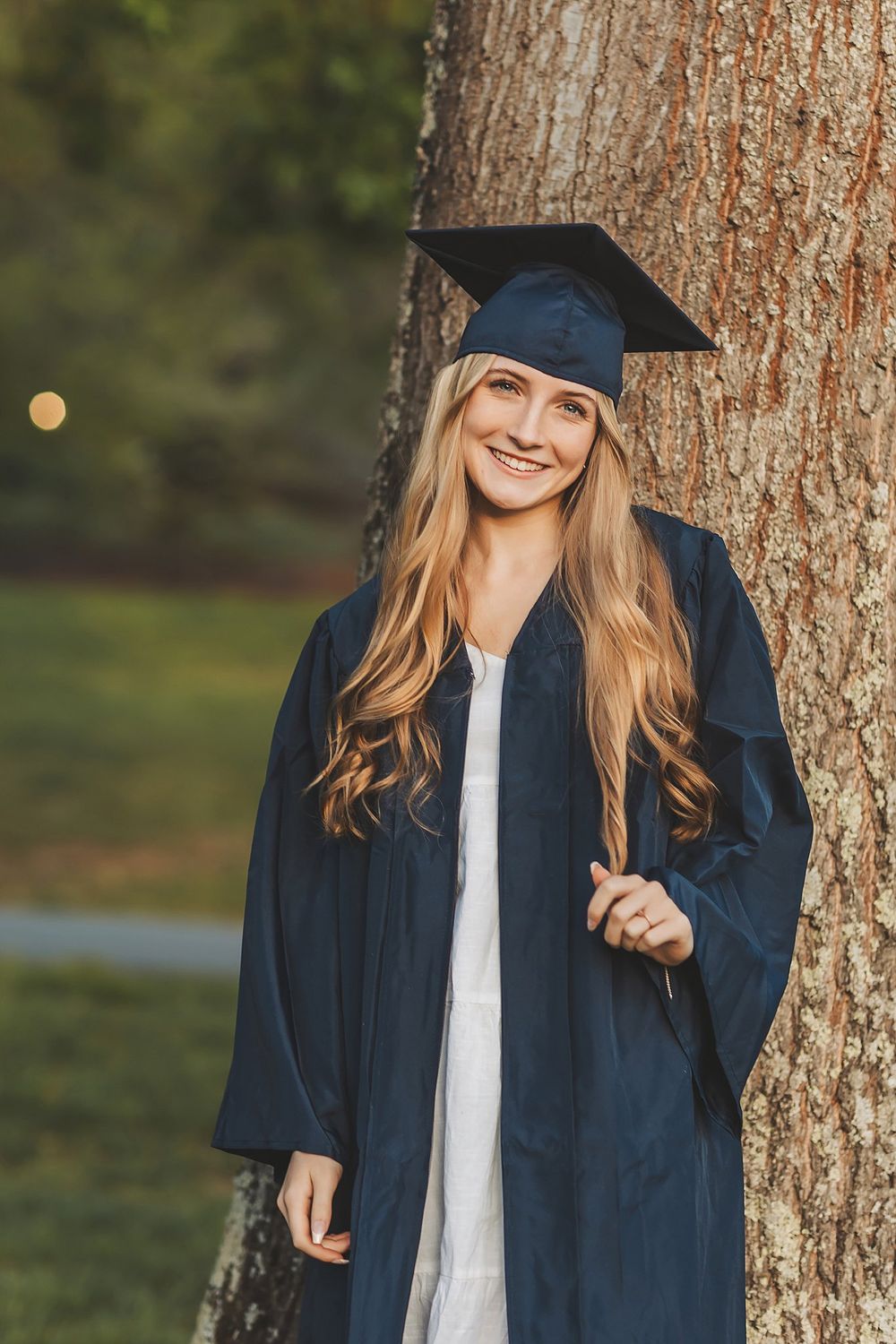 Graduate wearing navy blue cap and gown poses against tree trunk in outdoor setting.