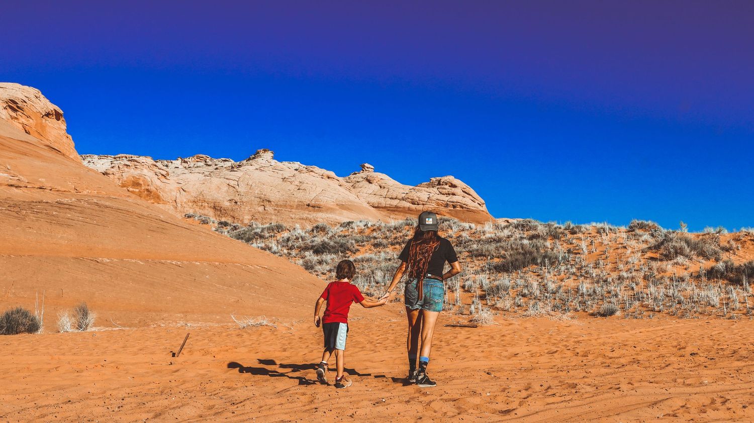 Adult and child walking hand in hand through a red rock desert landscape under blue sky.