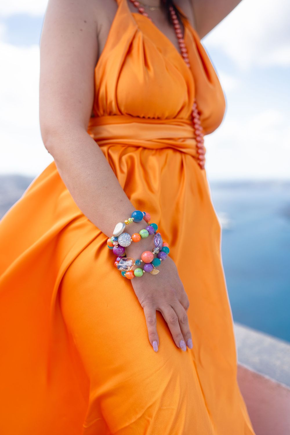 Orange halter dress detail with colorful beaded bracelet against ocean backdrop.
