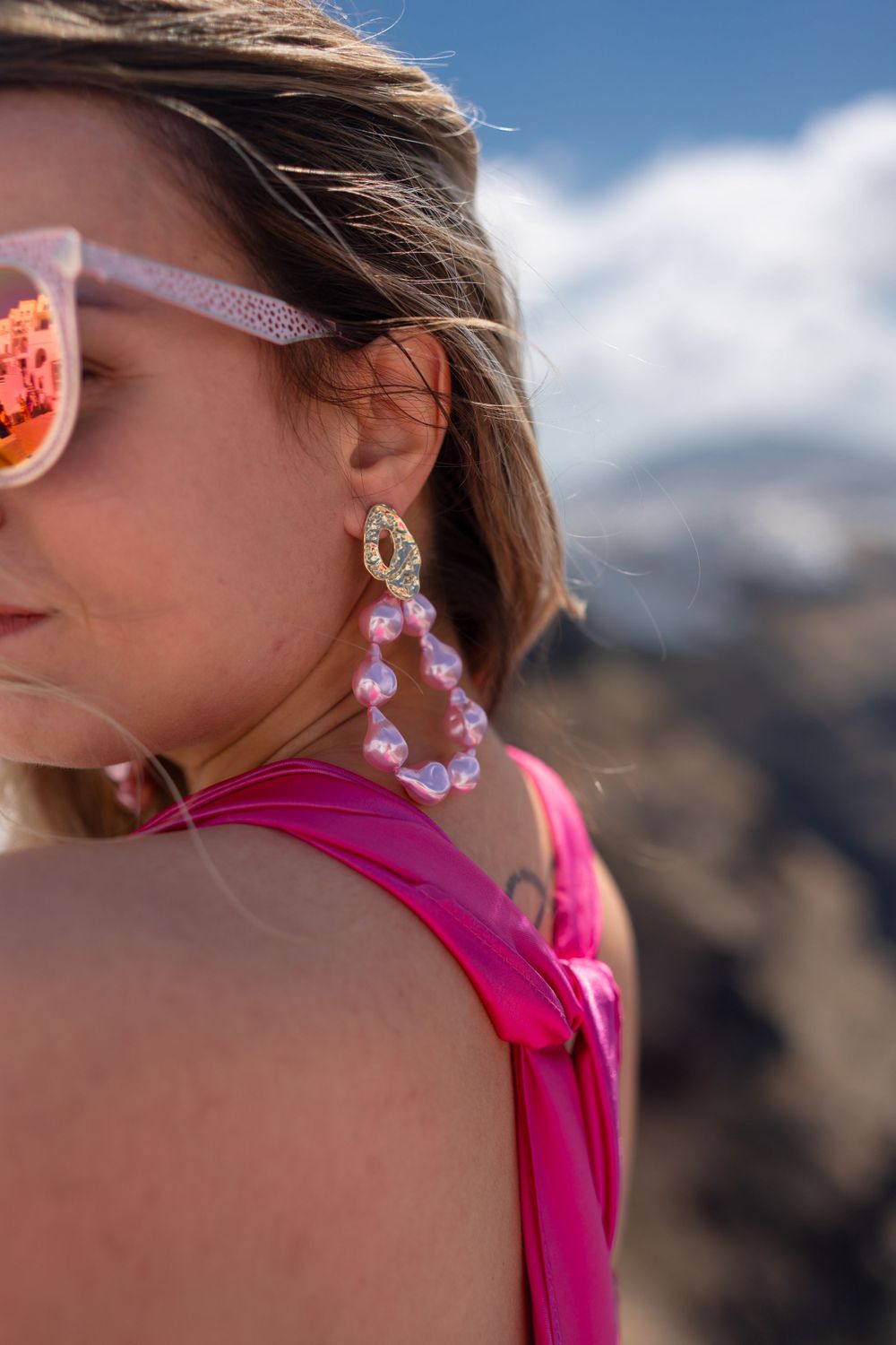 Close-up of pink sunglasses and pearl earrings against mountain landscape.