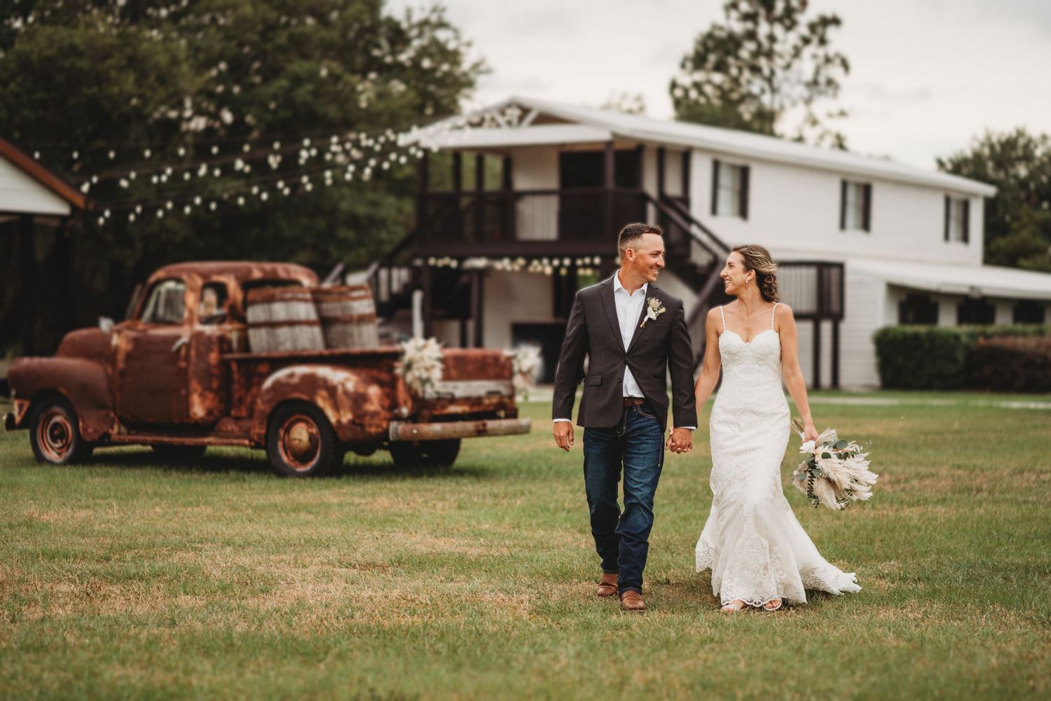 A couple walks hand in hand through a field with a vintage rusty truck in the background.