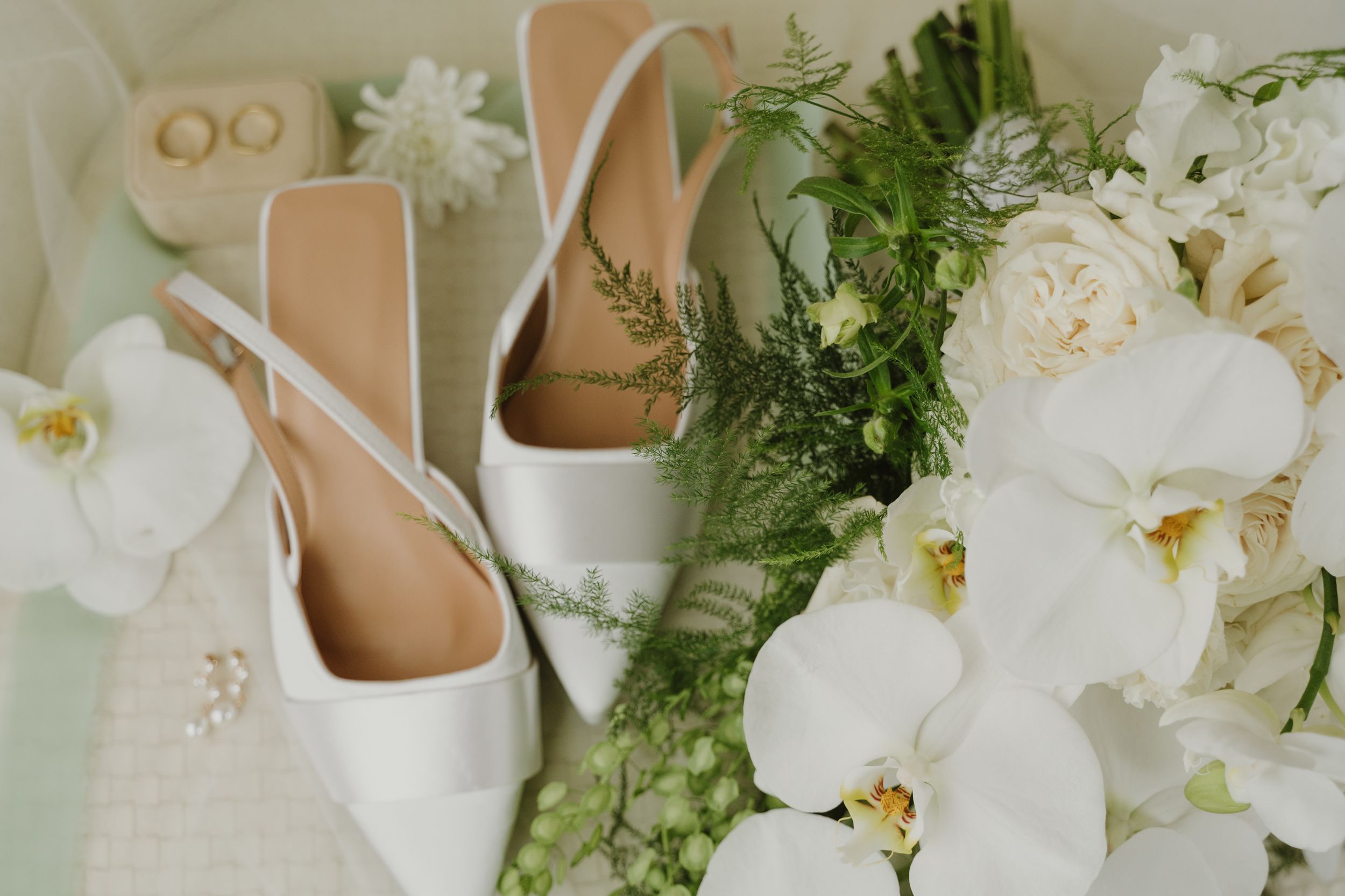 White bridal shoes and orchids arranged with wedding rings and greenery.