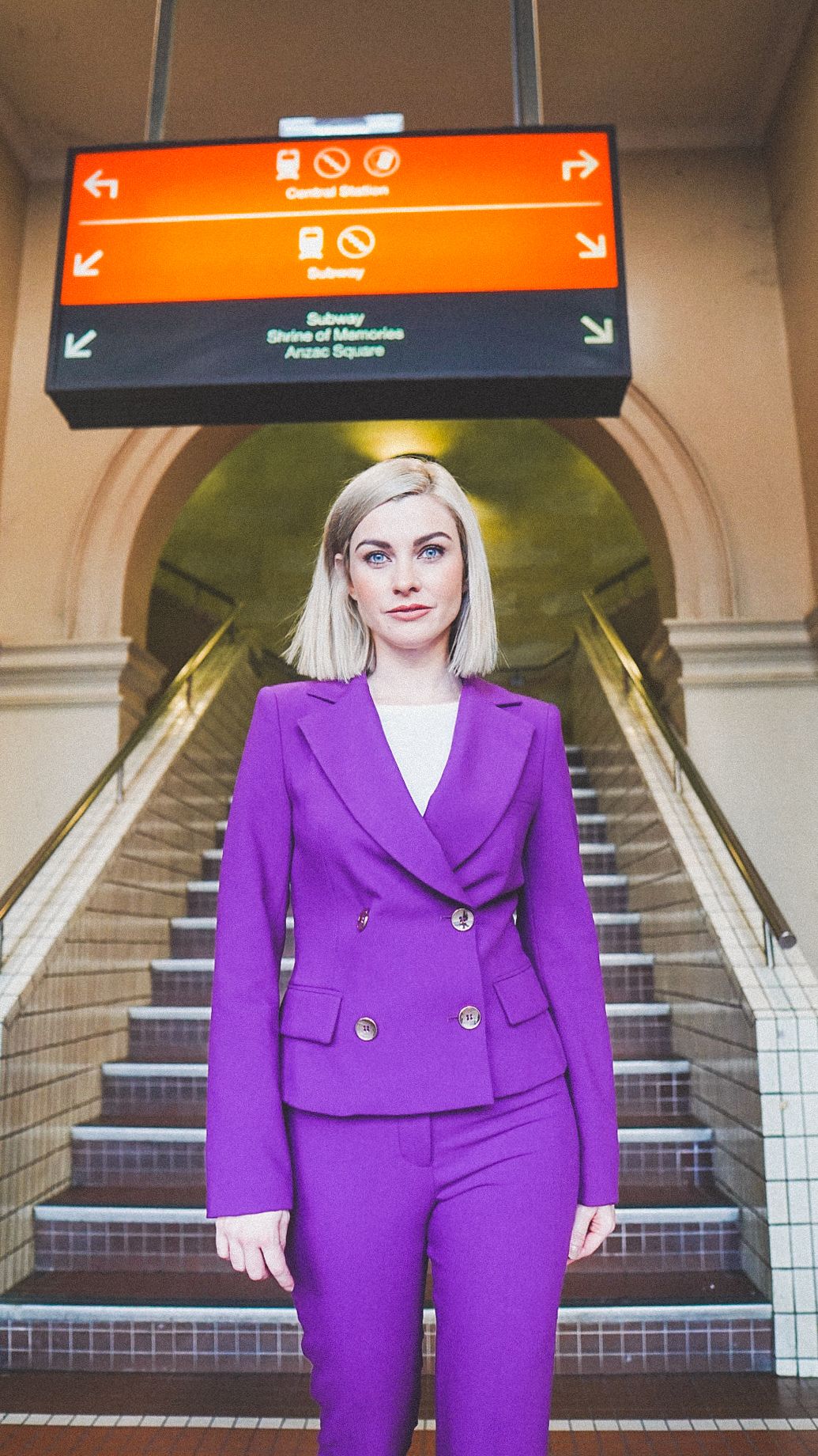 A stylish blonde woman wearing a vibrant purple suit poses in front of stairs at a subway station.