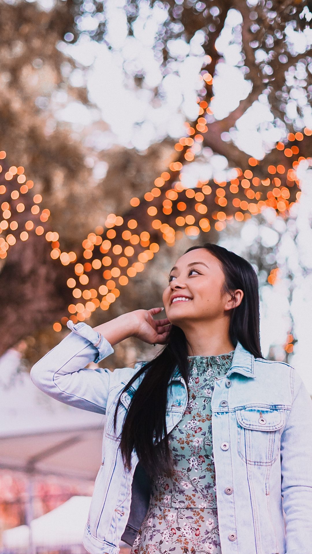 Happy young woman in denim jacket smiling under bokeh string lights in trees.