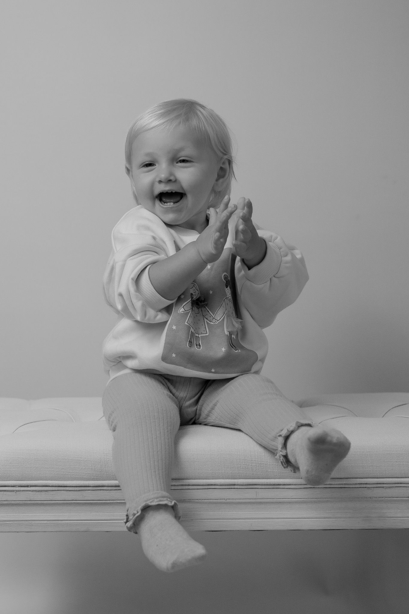 Black and white photo of a toddler clapping excitedly while sitting on a cushioned surface.