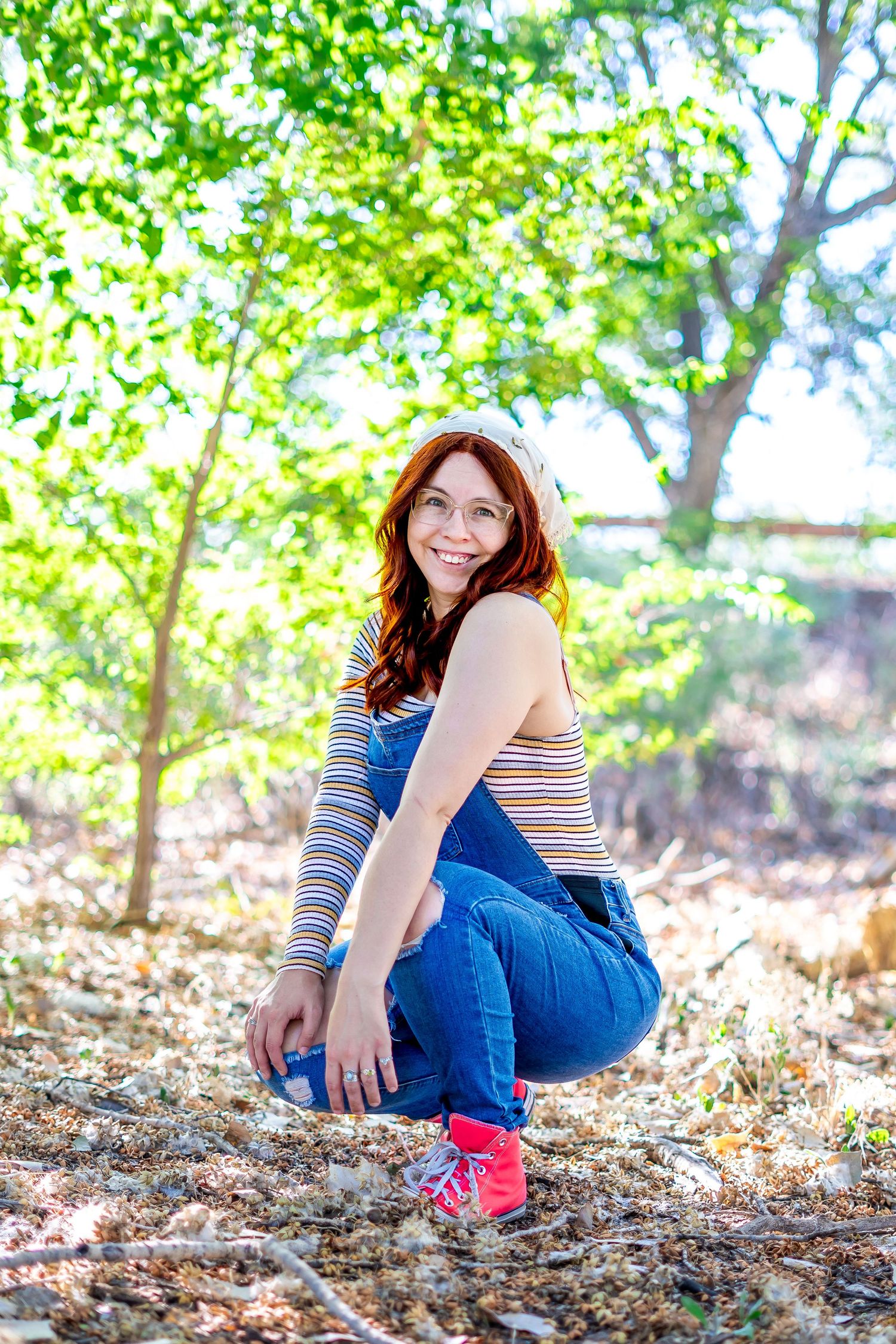Person in striped top and jeans crouches outdoors among green foliage and trees on a sunny day.