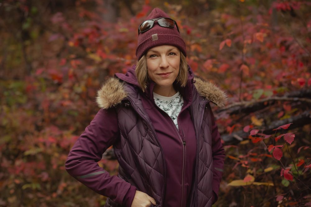 Alison of Lumenali Photography stands amongst the fall flowers wearing a maroon hat, vest and jacket, at Parrot Bay, Kingston, Ontario.