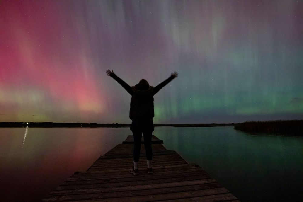 Alison silhouette stands on a floating dock, hands raised as northern lights, dance around her, reflecting off the lake. Camden Lake, Ont.