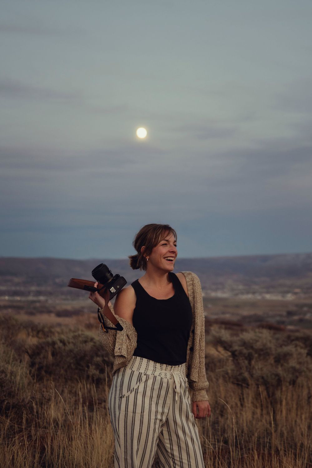 Ali of Lumenali Photography stands on top of a small mountain, holding her camera, moon rising in the background. Salt Lake City, Utah.